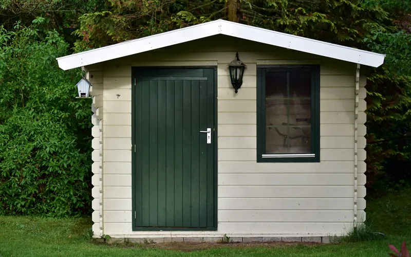 Abri de jardin en bois clair avec porte et fenêtre vertes, posé sur une pelouse devant une haie